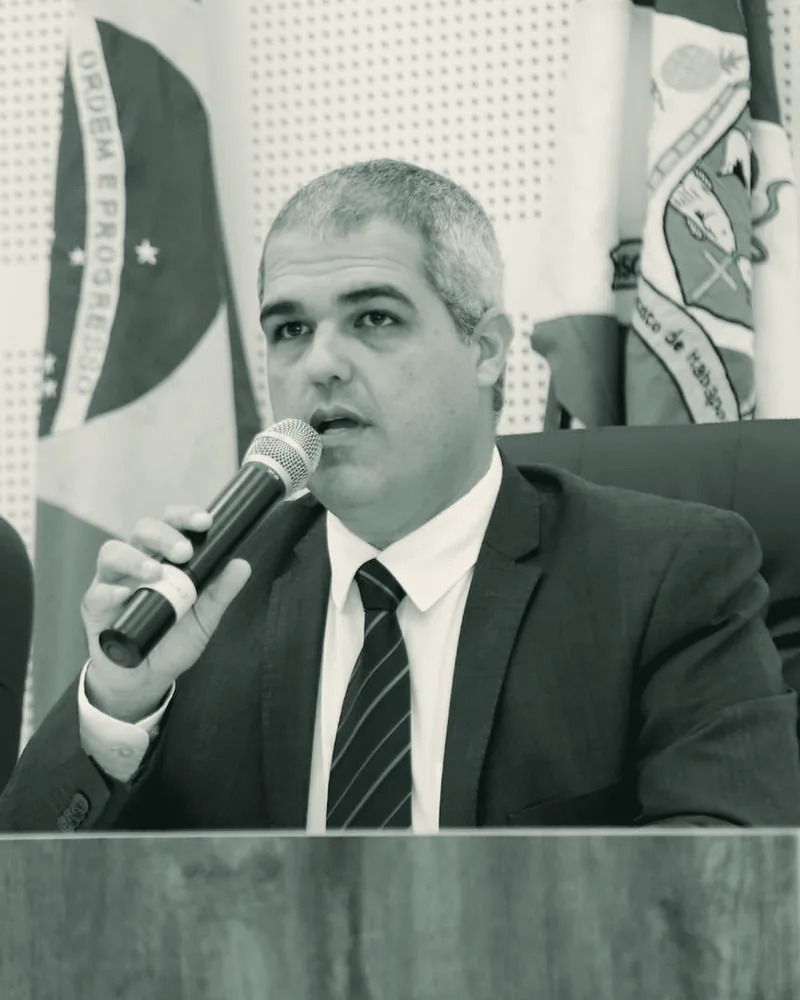 Fredy Beshara fala ao microfone na tribuna, com a bandeira de São Francisco de Itabapoana ao fundo